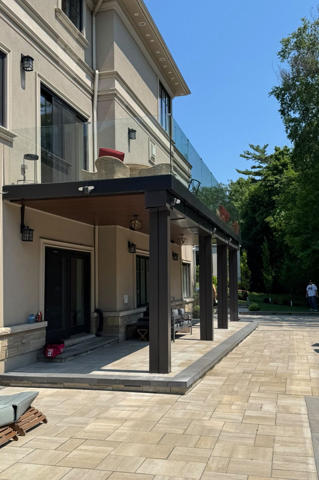 Covered patio with dark-clad beams and columns, a wood-look soffit and a frameless glass railing on the balcony above