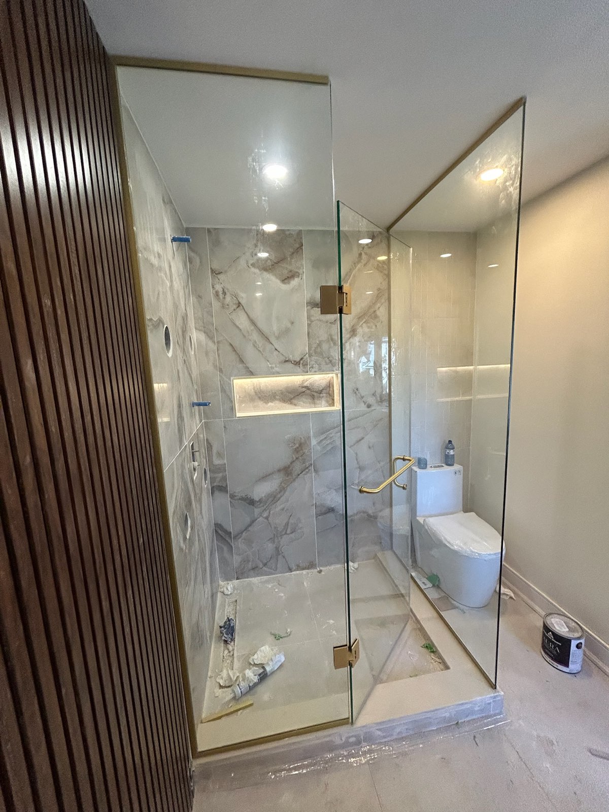 Frameless glass shower next to a dark wood slat wall and marble-look tile